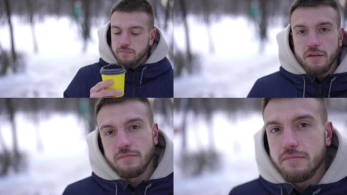 年轻的白人男子站在白雪皑皑的冬季公园的右边，喝着咖啡，看着相机。严肃自信的积极人物在户外慢动作摆姿势
