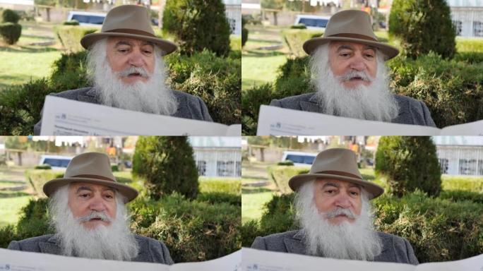 肖像一位聪明而时尚的老年绅士，留着美丽的胡须，胡须和眉毛，正在公园里的长凳上读报纸，背后是绿色美丽的