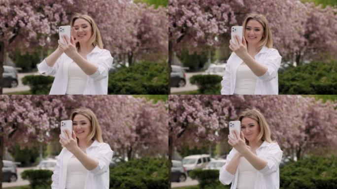 美女可以用智能手机视频通话。女孩站在樱花树前与鲜花交谈和微笑。春天晴朗的一天。