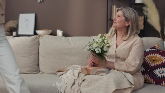 一位老年妇女在沙发上放松时翻阅相册。一个女儿带着一束鲜花拜访妈妈，庆祝母亲节，生日。高级女性的惊喜