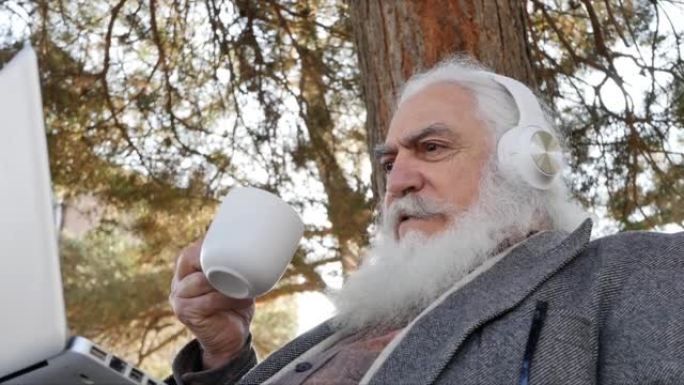 像圣诞老人一样有着美丽白胡子的野蛮优雅的高级男子正在用笔记本电脑和白色耳机进行视频通话，在公园里的长