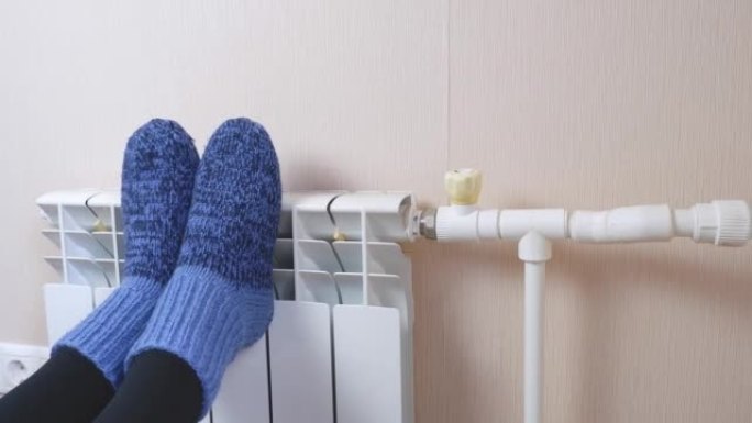 在寒冷的冬日，一名妇女穿着蓝色羊毛袜在散热器上温暖脚。中央供暖系统。寒冷季节昂贵的取暖费用