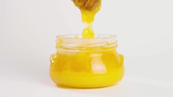 蜂蜜用蜂蜡从蜂蜜勺子里滴在白色背景的罐子里。天然液体蜂蜜从滴头流下来。