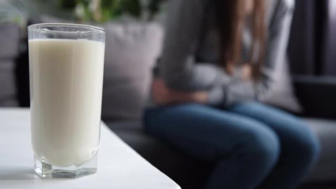 玻璃牛奶的选择性焦点是在白色的桌子上，不健康的年轻女性触摸胃患有严重的疼痛坐在家里的客厅沙发上。乳糖