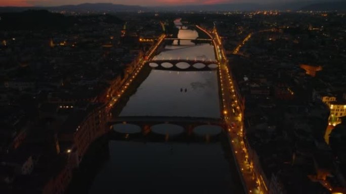黄昏时向后飞过阿诺河上。由路灯点亮的滨水街道和桥梁。向上倾斜显示五彩缤纷的天空。意大利佛罗伦萨