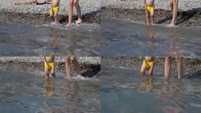 母亲的特写镜头，帮助2岁的儿子穿着黄色泳裤，带着鹅卵石进入海滩上的冰冷海水。暑假，假期，家庭旅行。没