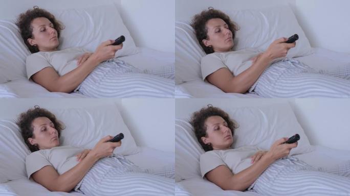 床上一个无聊的女人手里拿着电视遥控器。一头卷发的白人妇女穿着普通衣服躺在床上，看电视，用遥控器切换频