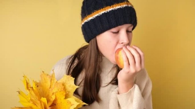 十几岁的女孩吃一个孤立在黄色上的苹果。穿着针织舒适衣服的可爱的年轻女孩在彩色工作室背景上咬苹果。健康