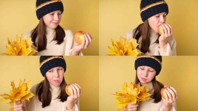 十几岁的女孩吃一个孤立在黄色上的苹果。穿着针织舒适衣服的可爱的年轻女孩在彩色工作室背景上咬苹果。健康