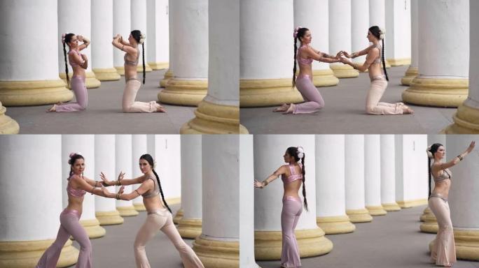两个才华横溢的漂亮女人跪在地上站起来，在城市的白色大柱子上表演印度舞蹈。自信美丽的表演者在户外跳舞的