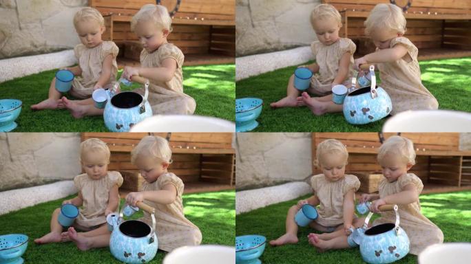 可爱的金发双胞胎姐妹正在操场上玩一个玩具厨房，穿着相配的衣服坐在草地上。游戏中的孩子模仿喝茶。
