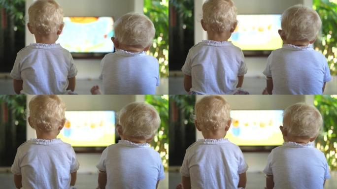 两个金发小女孩坐在一个大房间里，在大屏幕平板电视上看自己喜欢的动画片。儿童消遣的概念。