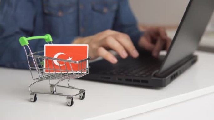 桌子上有土耳其国旗的小购物车。男子使用笔记本电脑在网上购物。