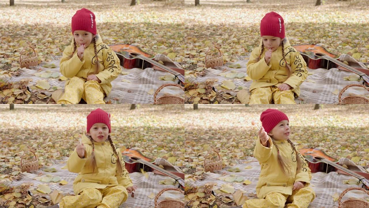 小开朗的学龄前儿童女孩温暖的红帽子吃舔棒上的大糖果。孩子在外面吃糖果野餐。秋天公园篮子里寒冷的天气里