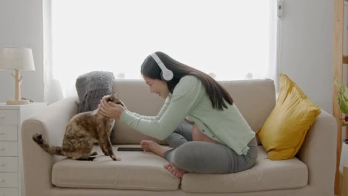 亚洲孕妇在家坐在沙发上玩猫，戴着耳机听音乐。怀孕、生育、准备和期望概念。