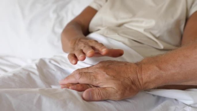 一个年迈的男人把手放在一个躺在床上的老妇人的手上。因此，他鼓励她，并在她长期治疗和住院期间给予她道义