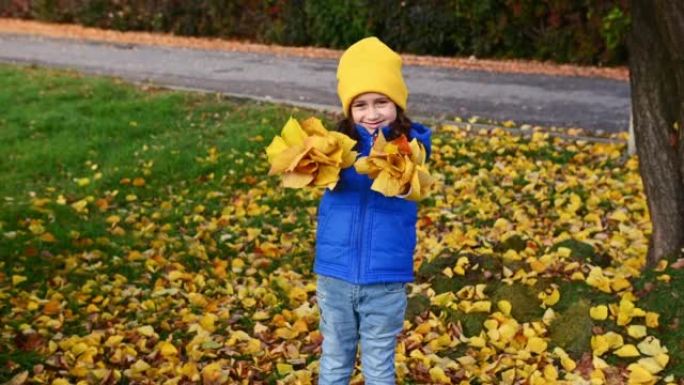 一个穿着蓝色夹克，黄色帽子和毛衣的小女孩，在公园散步时抛出了收集的秋天落叶