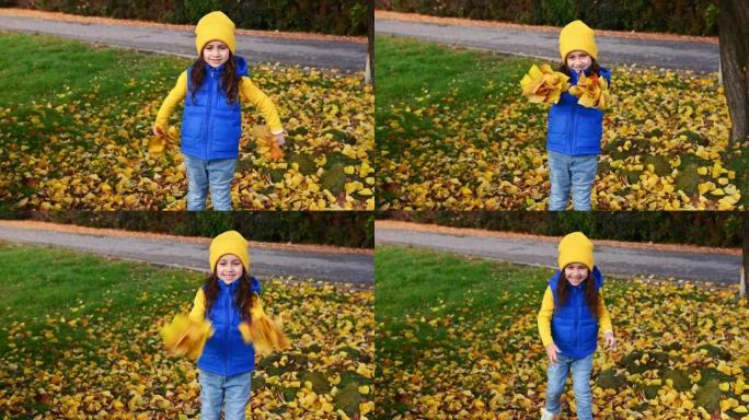 一个穿着蓝色夹克，黄色帽子和毛衣的小女孩，在公园散步时抛出了收集的秋天落叶
