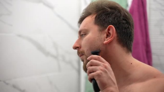 英俊的男人在浴室里用电动修剪器剃光脸颊、胡子和胡须，背景是彩色毛巾，年轻的男性在家做日常皮肤护理