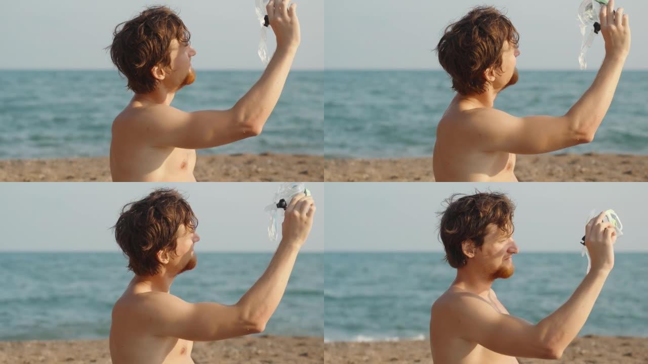 海滩上留着红色胡须的男人拿着水肺护目镜，看着太阳。大海在背景中。慢mo