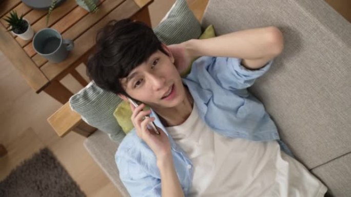从一个休闲的亚洲人的顶部看，他斜躺在沙发垫上，一边在家里的客厅里用手势在手机上聊天。