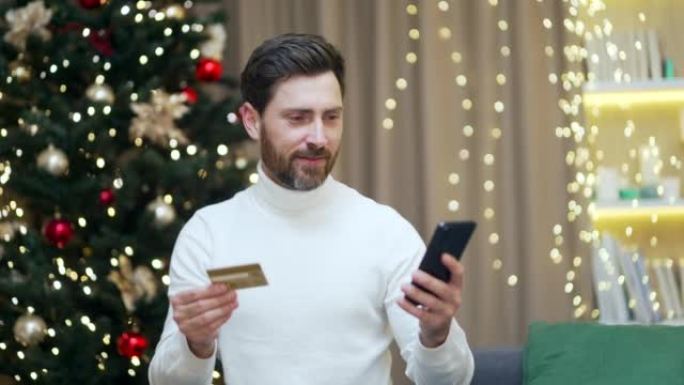 快乐男人在新年圣诞节假期在家中使用手机和信用银行卡在线购物