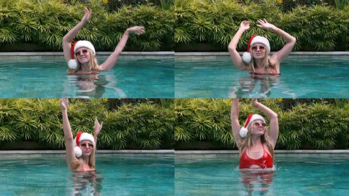 快乐的年轻金发女子在度假时在游泳池游泳时戴着圣诞老人帽子、aed比基尼和太阳镜。女孩举起双手跳舞，在