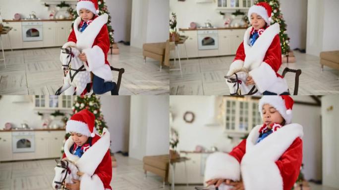 穿着蓝色圣诞毛衣和红色圣诞老人帽子的非洲裔美国小女孩，坐在圣诞树和节日餐桌的背景下，坐在玩具摇马上微