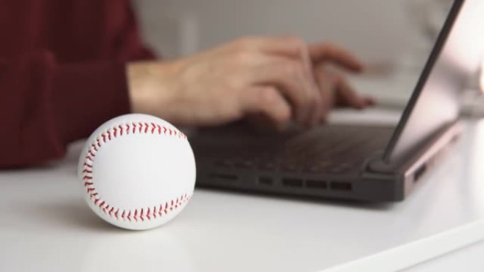 年轻人检查棒球统计数据或在笔记本电脑上写棒球文章。桌子上的白色皮球。博彩，赌博的概念。
