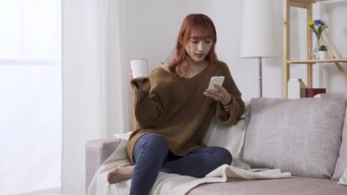 在家里的沙发上放松，冬天的漂亮女人穿着休闲服，喝着早晨的咖啡，在智能手机上浏览在线新闻。真正的生活方