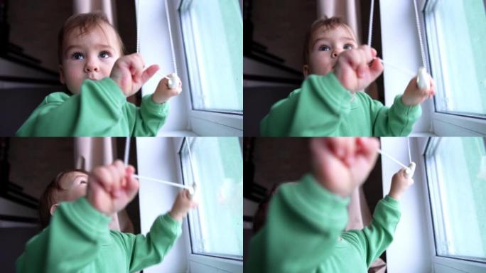 好奇的活跃的高加索孩子从jalousie窗帘上拉着绳子。男婴站在窗前玩耍，兴致地抬头。特写。
