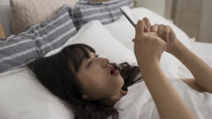 惊讶的亚洲妇女在家里的卧室里使用智能手机躺在床上时，听到一个令人震惊的在线新闻，她张开嘴。