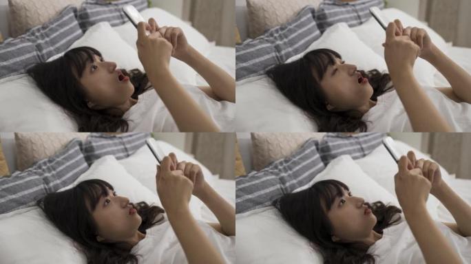 惊讶的亚洲妇女在家里的卧室里使用智能手机躺在床上时，听到一个令人震惊的在线新闻，她张开嘴。