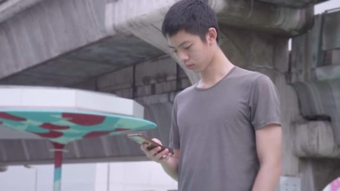 一个年轻的亚洲男孩，高个子，聪明，英俊，穿着t恤，牛仔裤，站着玩他的智能手机