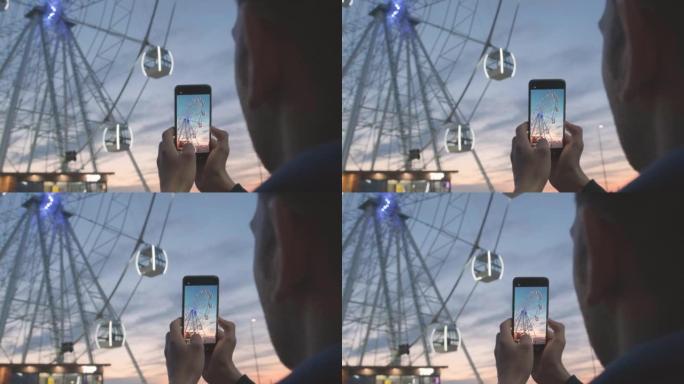 男子在日落时使用手机上的摄像头制作摩天轮视频。在智能手机上拍摄狂欢节的吸引力