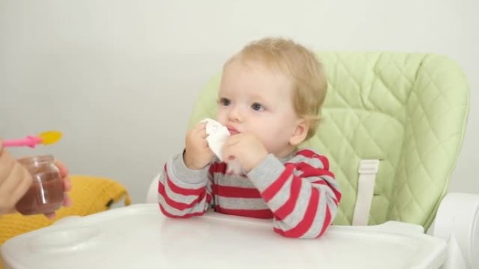 有趣的女婴用餐巾纸擦嘴，坐在高脚椅上，妈妈喂孩子，用勺子做蔬菜午餐，婴儿断奶，给小孩子的第一份固体食