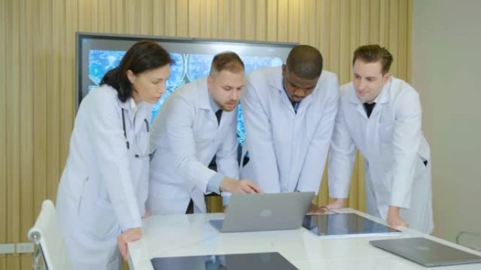 医生团队站在医院会议室，主任医师讨论在笔记本电脑中寻找患者的治疗方法。穿着白大褂的同事一起分析疾病症