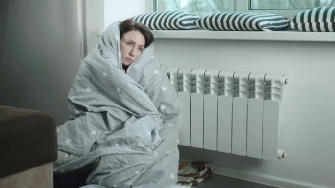 欧洲妇女裹着毯子，试图在散热器附近保暖。能源危机，节约住宅热量。
