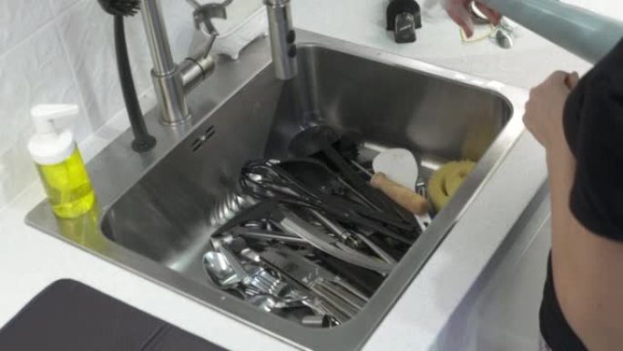 女人用海绵叉子洗勺子在厨房水槽里水流下，女人用手洗餐具特写。