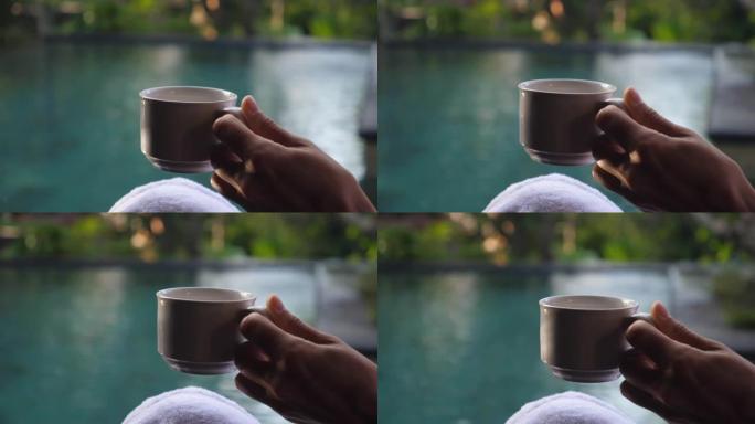 女人的手在私人碧绿的水池的背景下握着一杯咖啡。早晨的阳光在杯子上播放。完美的早晨概念。