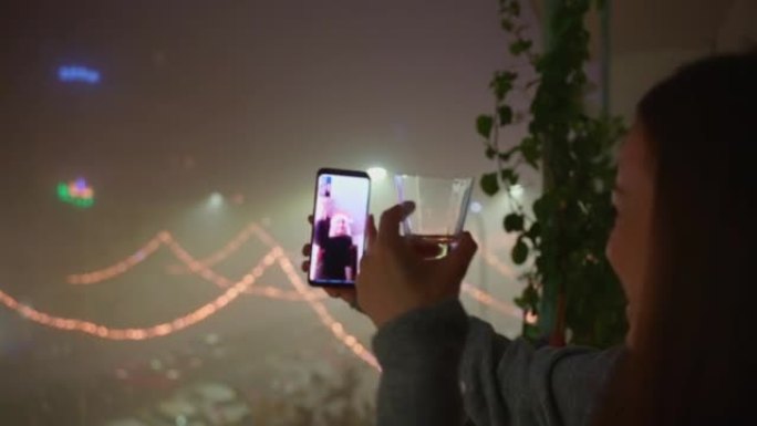 快乐微笑的女人，手里拿着一杯香槟，通过与家人或朋友的视频通话，在远处欢呼，女人站在阳台上，在雾中展示