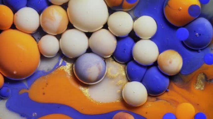 特写多色的蓝色橙色和白色气泡在宇宙色彩表面破裂并混合在抽象设计中。金色闪闪发光的颗粒、墨球滴和混合