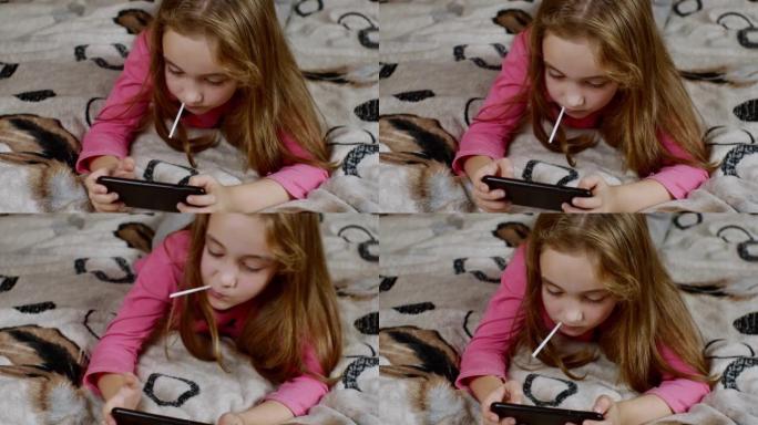 一个可爱的小女孩躺在沙发上，吮吸着甜美多汁的棒棒糖，在智能手机上热情地玩耍，并在胜利中举手。青少年数