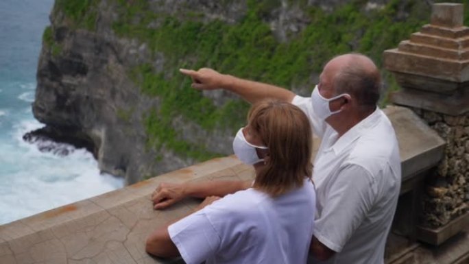 老年夫妇在观景台上戴着防护口罩，欣赏着令人惊叹的海景。在大流行期间旅行。新型冠状病毒肺炎概念。