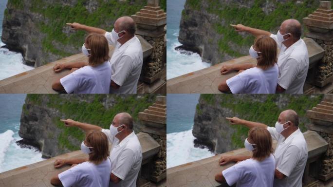 老年夫妇在观景台上戴着防护口罩，欣赏着令人惊叹的海景。在大流行期间旅行。新型冠状病毒肺炎概念。