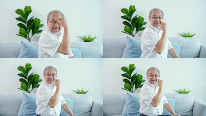 亚洲高级祖父的肖像坐在沙发上，做着振作的手势或身体健康。