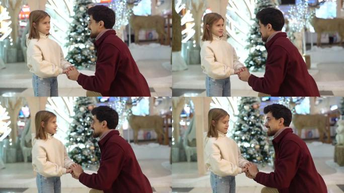 在美丽的圣诞节装饰背景下，欢快的爱心爸爸站在膝盖上与小可爱女儿在购物中心交谈的侧视图。