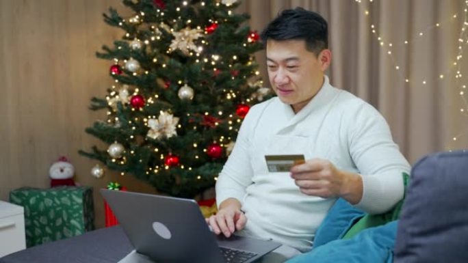 亚洲男子使用笔记本电脑和信用银行卡在线购物，在新年圣诞树冬季假期坐在家里的沙发上