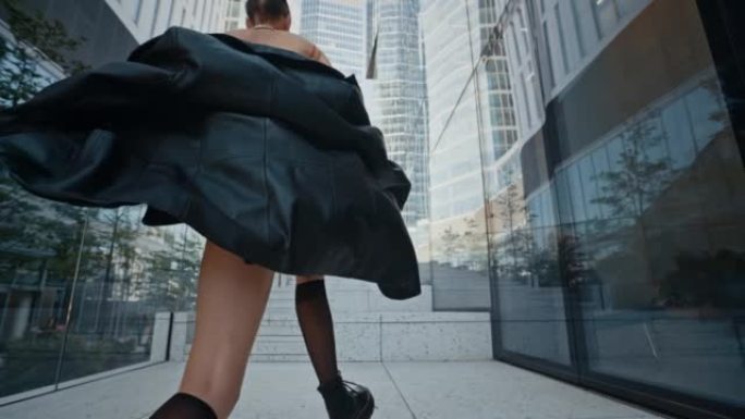穿着黑色皮夹克和短裙的时尚女孩在现代城市中心的高层玻璃建筑旁边爬上楼梯。时尚女性，夏日，慢动作