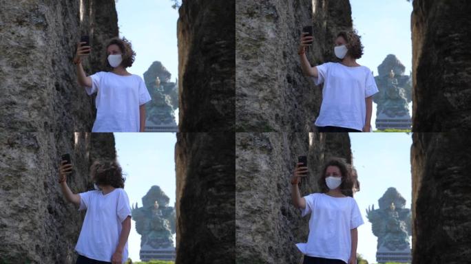 独自去异国他乡旅行。一名戴着病毒面具的年轻女子站在粪便之间，在巴厘岛神庙前自拍，然后回头。